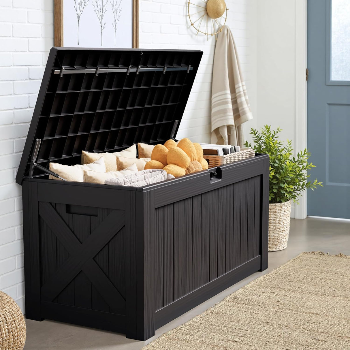 Patioslife Outdoor Resin Deck Box with open lid storing pillows and blankets in a covered entryway with white brick wall and blue door