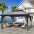 Patioslife Haven 10' x 12' grey hardtop pergola gazebo with double vented roof and side curtains, set on a stone patio beside a coastal home with ocean view and outdoor sofa seating