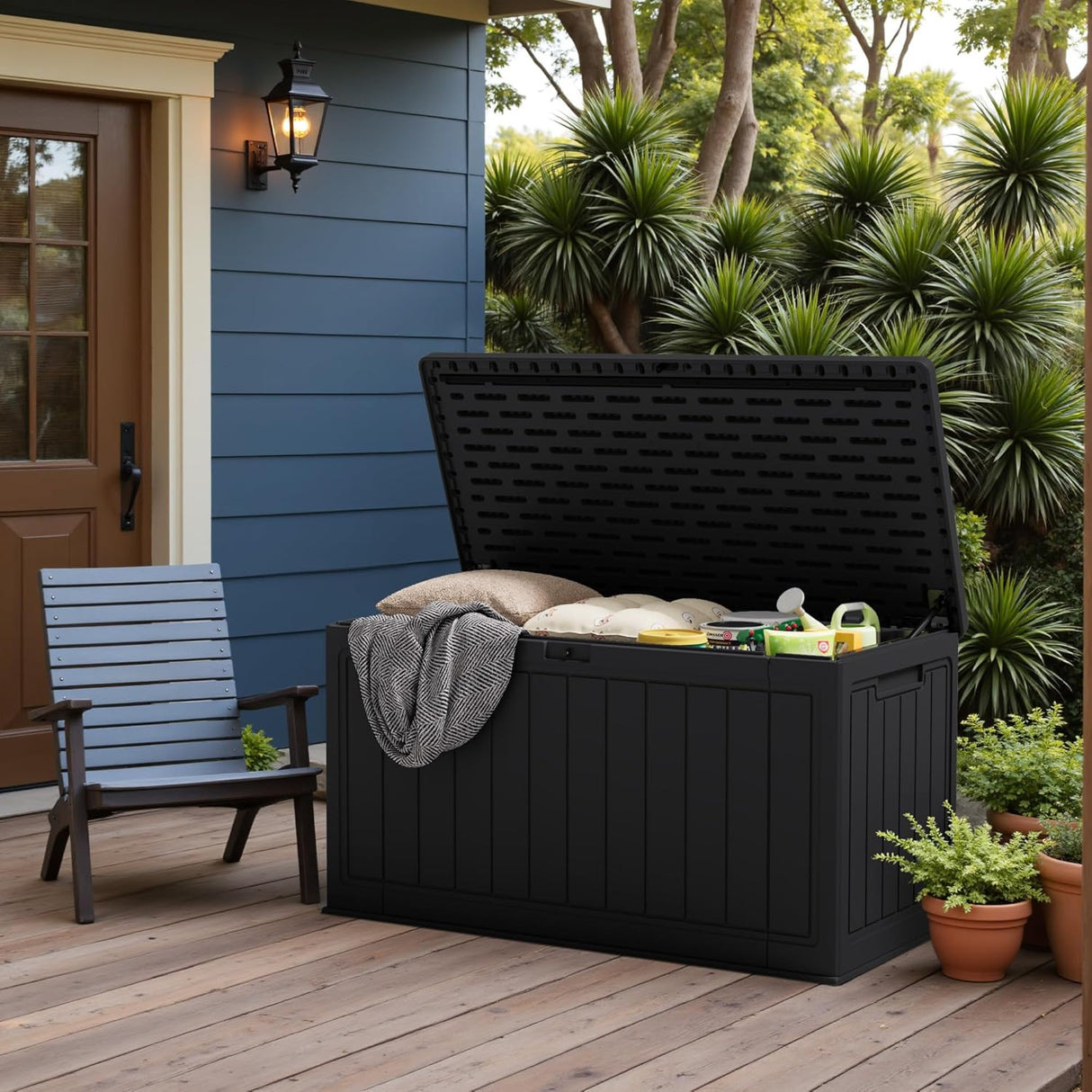 Patioslife Outdoor Resin Deck Box open on a wooden porch beside a blue house, storing patio pillows and outdoor supplies near garden plants